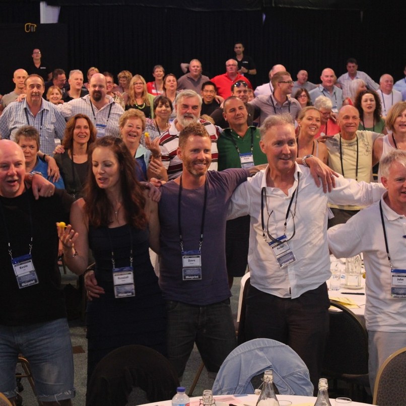 A room of people with their arms around each other, singing and swaying together at an Amanda Gore keynote